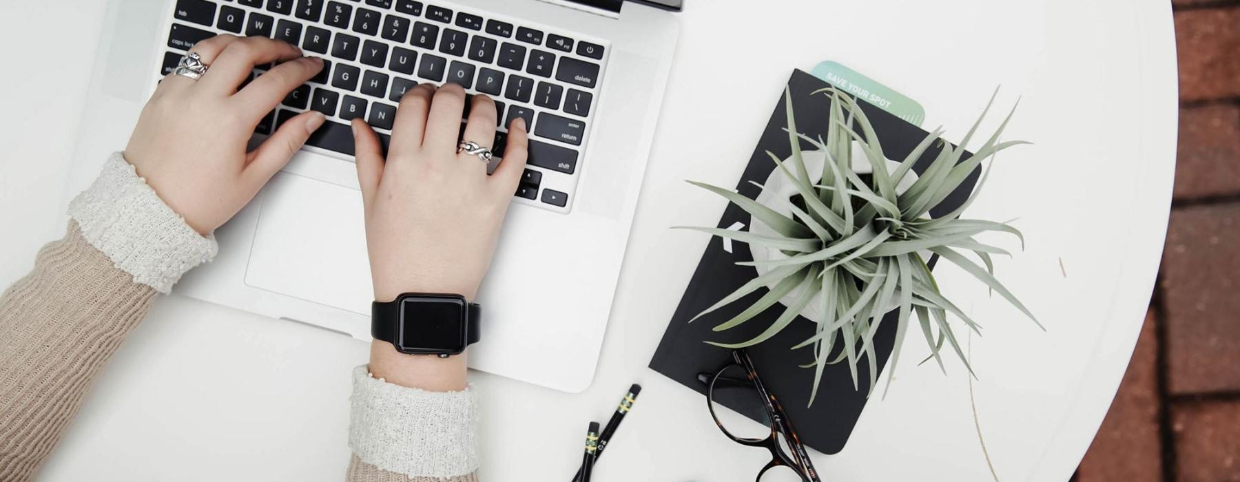 overhead shot of woman working on a laptop at a round table with pencils, her cell phone, glasses, notebook and a potted plant